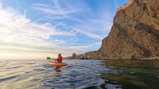 Kayaking sunset ocean man paddles orange kayak along high rocky cliffs during beautiful golden hour for peaceful outdoor travel adventure