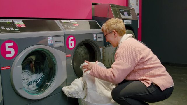 Woman with short blonde hair wearing a pink sweater loads laundry into washing machine at selfservice laundry facility with modern appliances and bright decor