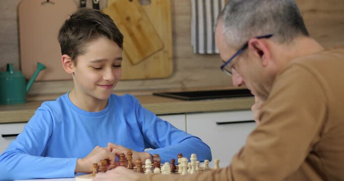 Smart child makes a chess move and smiles. Boy looks at board with excitement. Father and son play strategy game in kitchen. Kid is winning the match. Intellectual hobby is fun. Intellectual family.
