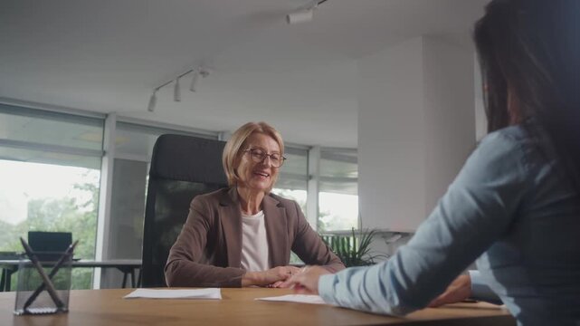 Medium shot of businesswoman in suit showing where to sign document to brunete woman at desk in her office