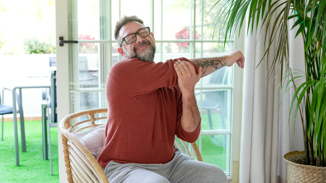 Active mature bearded man exercising inside home with stretching exercises for shoulders and arms in a modern living room. Healthy  lifestyle, strength training, and wellness concept