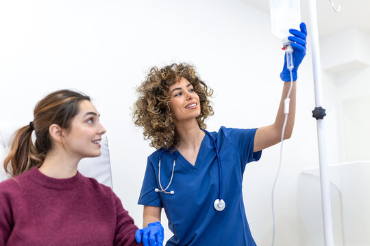 A curly-haired doctor in blue scrubs looks up while adjusting a clear IV infusion bag on a stand. A young female patient sits nearby. Concept of medical care, hydration therapy, and hospital service.