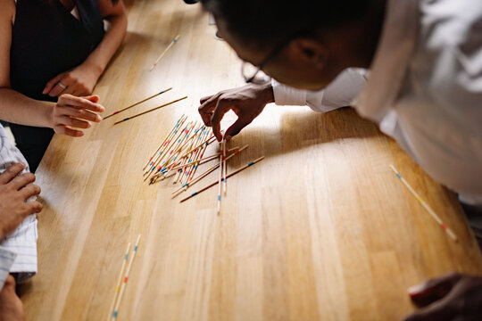 Friends gather around a warm wooden table, laughing and focused, as they carefully navigate a colorful array of pick-up sticks, creating a joyful atmosphere of competition and fun.