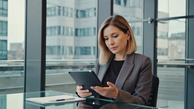 Middle-aged European female chief operations officer reviewing quarterly operations data on tablet in modern office with large windows and city view