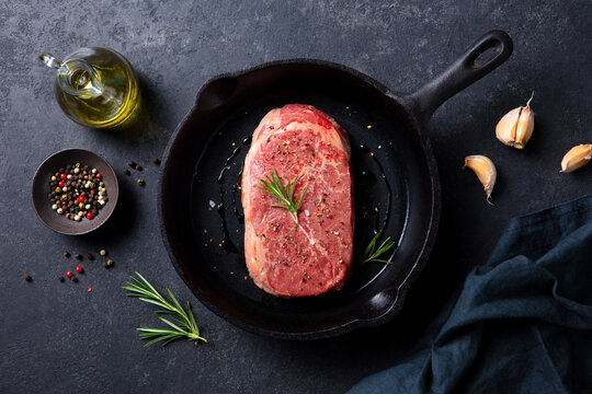 Fresh raw steak in cast iron pan, with rosemary touch, garlic pieces, peppercorns scattered, olive oil accent, dark aesthetic. Grey background. Top view.