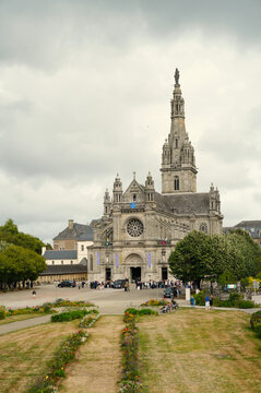 View of the Basilica of Sainte-Anne d'Auray in Brittany, France. The stone neo-Gothic facade features a tall central tower, a large rose window, and ornate sculptures under a dramatic cloudy sky