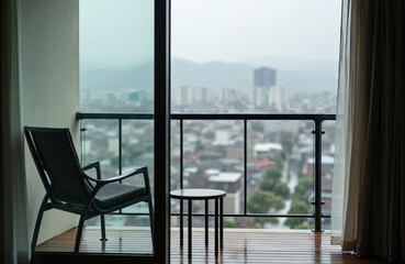 Naklejka premium View from apartment window on rainy city with balcony chair and table. Modern buildings and distant mountains in fog. Overcast sky, grey weather. Urban landscape.