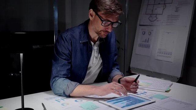 Professional man working late in a dark office, analyzing business data reports and using a digital tablet, managing overtime and workload under focused concentration