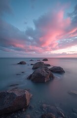 Rocky sea coast with gentle waves at sunset. Sky fills with soft pink and blue clouds. Long exposure creates dreamy water motion effect. Serene natural landscape.