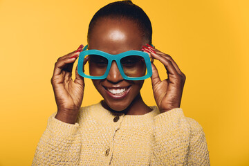 Smiling African woman with dark skin wearing large blue glasses and beige sweater stands against yellow background. Concept of happiness, fashion, and joyful lifestyle with bright colors. © SHOTPRIME STUDIO