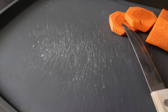 A kitchen knife cuts carrot pieces on a damaged plastic board with deep cut marks. Concept of microplastic contamination, unsafe kitchen tools, environmental impact, material degradation.