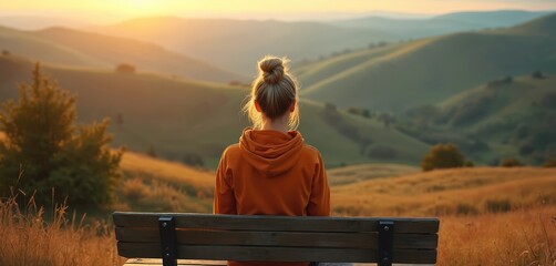 Naklejka premium Woman sits on bench watching golden sunset over rolling hills. Enjoying peaceful view on vacation trip, feeling calm, serene nature landscape. Female rests after long hike.