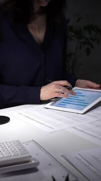 Woman working late night in office, multitasking with a tablet and writing on documents, managing heavy paperwork load. Audit and taxes in business