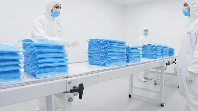 Two Asian women in white protective suits and blue masks organize stacks of blue surgical masks on a conveyor belt in a clean room environment with bright lighting
