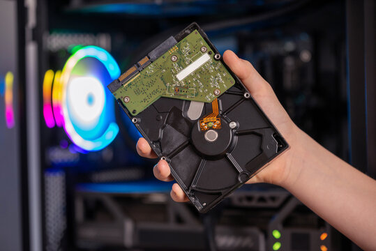 Hand holding opened hard disk drive in front of gaming computer with RGB lighting, internal storage component for data recovery and backup