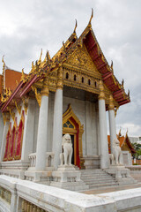 Fototapeta premium Closeup of the Marble Temple in Bangkok. The image showcases the white marble columns and the intricate golden gables of the multi-tiered roof. One of the white lion statues is in the foreground.