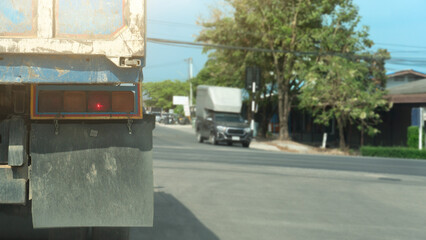 Rear view of a large truck with turn on brake light. Stop on the asphalt road in the junction. background of other car opposit road under green trees in the country. Under clear sky.