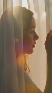 Serene woman gazing through sheer curtains in soft morning light in a tranquil environment from a close-up viewpoint
