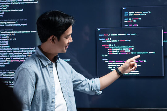 Asian Male developer leading a team meeting, pointing to code on a big monitor while reviewing an AI chatbot module. Concept of collaboration, debugging for coding teamwork in a tech company office.