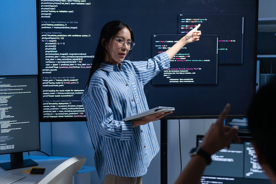 Asian Female Developer Leader pointing to monitor, presenting code on screen, explaining code to colleagues Male Developer coworker asking and discussing working together collaboration in office.