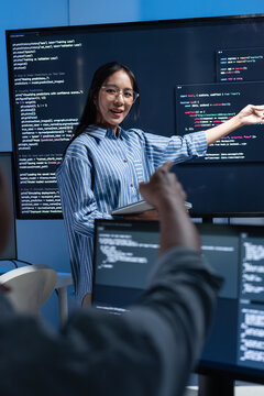 Asian female software engineer presenting source code on large screens in a modern tech office, explaining solutions in a team meeting. Concept for coding, code review, agile workflow.