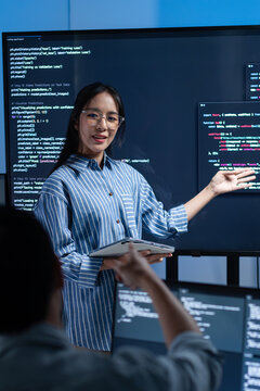 Asian Two Developer working on coding task, Collaboration in office. Female Programmer leader presenting code on monitor and discussing with team during software development meeting in offie at night.