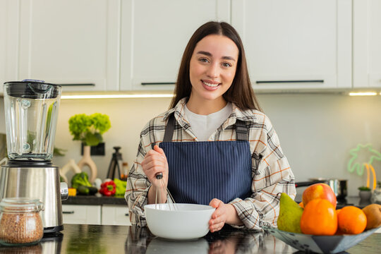 Young woman mixing batter with whisk in home kitchen for homemade meal prep healthy nutrition goals. Girl smiling during cooking practice creating fresh recipe for family dinner in apartment indoors.