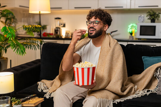 Arabian man at home watches movie on tv holds popcorn laughs comments enjoys calm evening. Egyptian guy on sofa streams show in blanket talks to screen smiles reacts to funny moments at night indoors.
