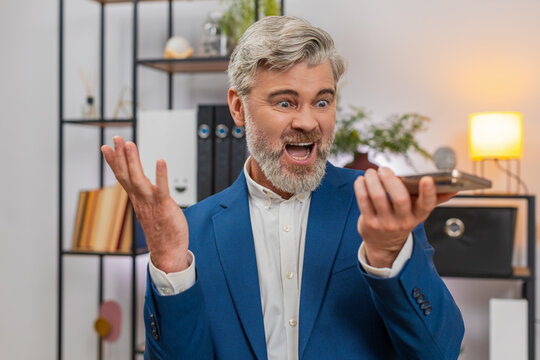 Middle-aged businessman at home office on smartphone speakerphone argues with supplier over shipment delay. Freelancer guy pounds table gestures angrily demanding delivery resolving client contract.