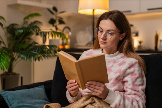 Middle-aged woman at night home wears glasses reads favorite book relax calm enjoys quiet time comfort. Caucasian girl on sofa at evening turns page smiles cozy mood peace rest gentle focus happy soft