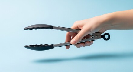 Obraz premium Hand holding black silicone kitchen tongs, gripping position, plain sky blue background.