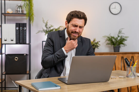 Middle-aged businessman at home office touches throat feels sharp pain when swallowing and keeps working on laptop. Freelancer guy at table sips water takes pill and searches help hoping for relief.