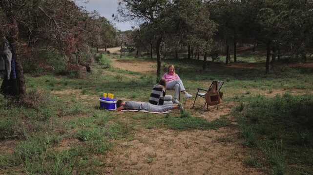 Group of three women relax outdoors in a grassy area, with one lying on a blanket, another seated in a chair, and the third sitting beside a cooler, surrounded by trees and nature