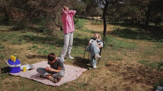 Group of three children enjoying a picnic in a grassy area, with one child sitting on a blanket, another standing, and a third relaxing in a chair, surrounded by trees and picnic items
