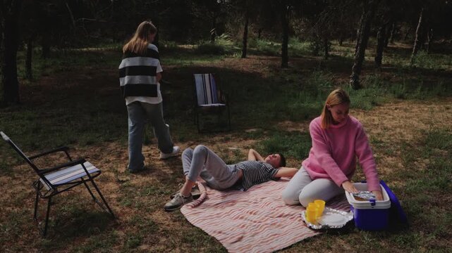 Group of three women enjoying a picnic in a forest, with one woman preparing food, another lying on a blanket, and the third standing nearby, surrounded by trees and outdoor seating