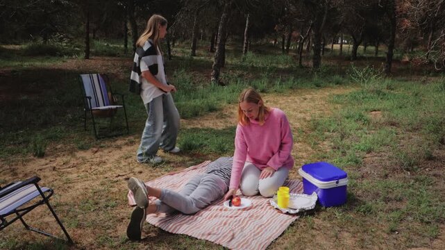 Two women in casual clothing interact in a natural setting, one preparing food while the other stands nearby, observing a third woman lying on a blanket in a serene outdoor environment