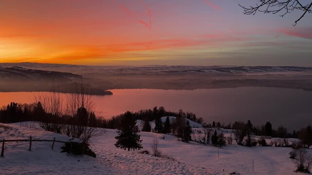 Intense winter sunset on Zugerberg overlooking Lake Zug, snow-covered landscape glowing under a dramatic red and golden evening sky.