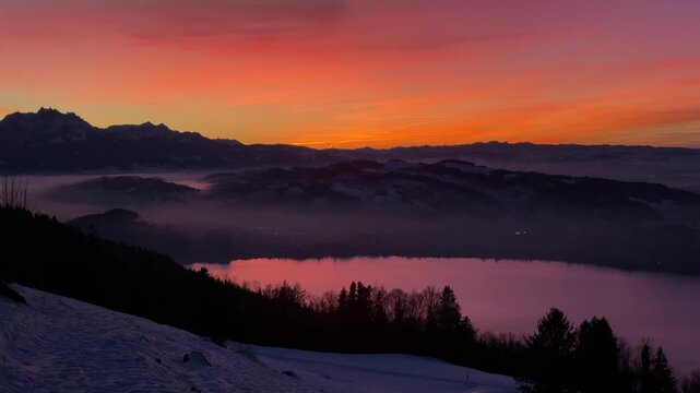Intense winter sunset on Zugerberg overlooking Lake Zug, snow-covered landscape glowing under a dramatic red and golden evening sky.