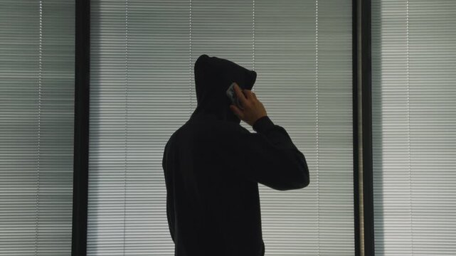 Silhouette of man with black hoodie talking on mobile phone against window. Suspicious spy or hacker walking and talking holding smartphone. Mysterious anonymous person inside apartment or home