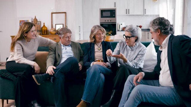 Five cheerful elderly friends gathering at home. They are sitting on a couch, talking and smiling together, enjoying a friendly conversation