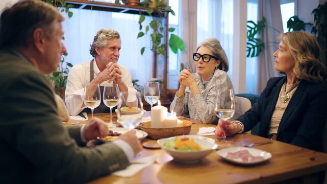 Group of elegant older friends talking and laughing together. Happy senior people having a pleasant dinner party in a cozy candlelit room