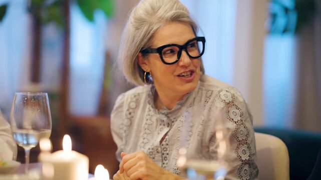 Cheerful senior woman with grey hair and glasses enjoying a candlelit dinner. She claps, smiles, and passionately tells a story to her friends
