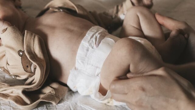 A close up shot of a person's hands checking and changing a disposable diaper on a small infant lying on a bed in a natural light setting.