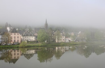 Fototapeta premium Berncastel Wehlen spiegelt sich im Fluss Mosel bei Nebel am frühen Morgen im Moseltal im Frühling