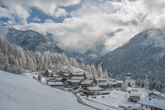 Snow-covered Bschlabs in Tyrol, Austria, nestled in the Lechtal Alps. Quiet alpine village in winter with deep snow, dramatic mountains, and soft sunlight breaking through mixed clouds.