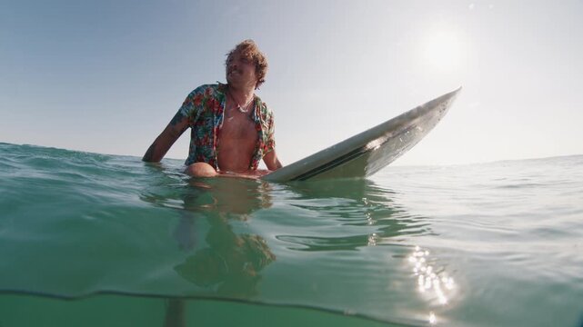 Hippie surfer. Young man in colorful stylish shirt sitting on surfboard in the ocean waiting for the next. Brazilian hippie surfer