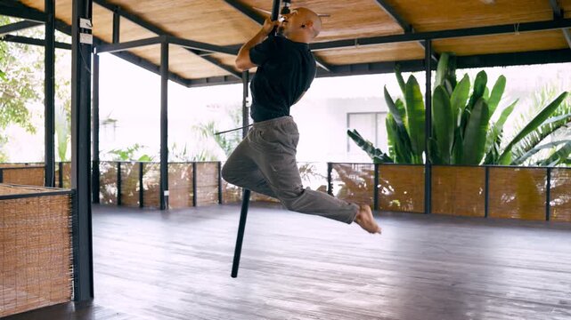 Athletic bald man demonstrating strength and flexibility while performing acrobatic elements on a pole in a bright, spacious dance studio