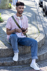 Fototapeta premium Asian man sitting on rounded curb holding small brown object while wearing headphones and green bag