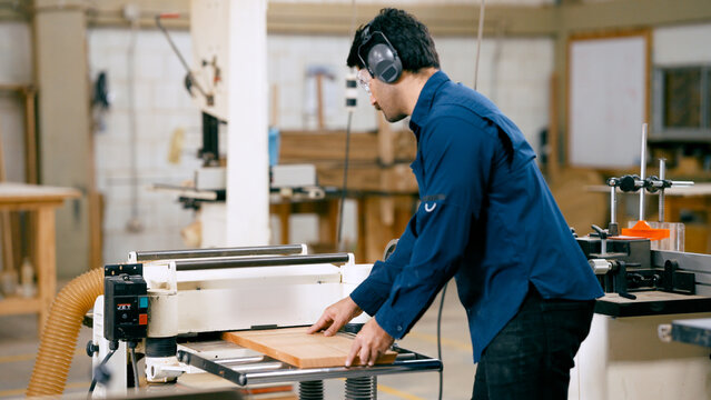 Skilled carpenter smoothing wooden board with a thickness planer, wearing safety gear in a woodworking shop
