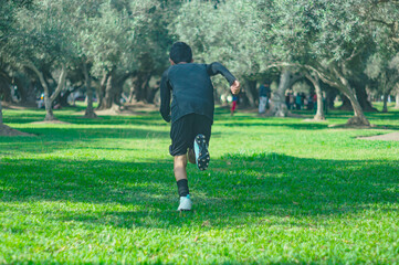 A unrecognizable young boy seen from behind in sportswear is captured in mid-stride, running enthusiastically through a park with a background of lush trees and dappled sunlight.